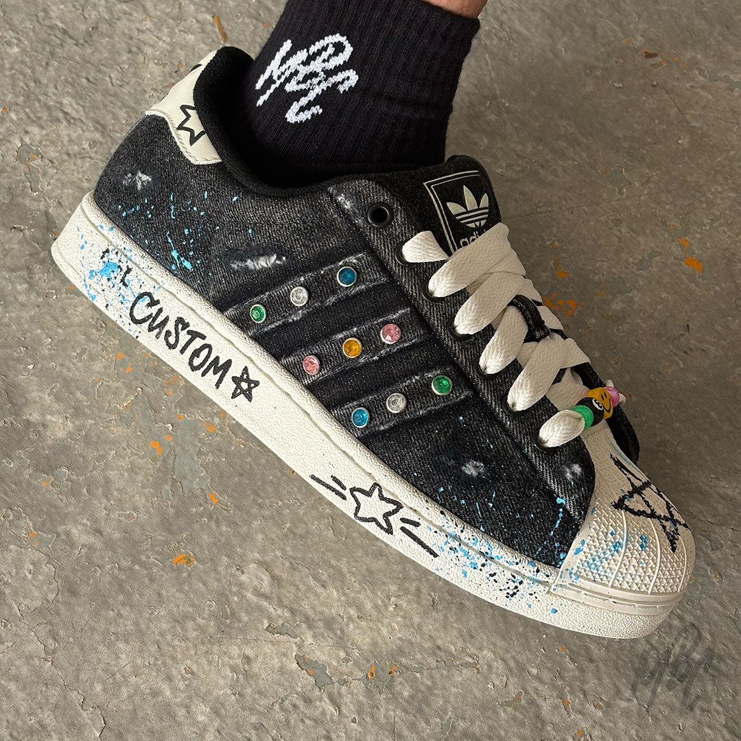 Stardust Denim custom Adidas Superstar worn on foot, featuring black denim uppers, colourful crystal details, white rope laces and hand-painted blue paint splatters.