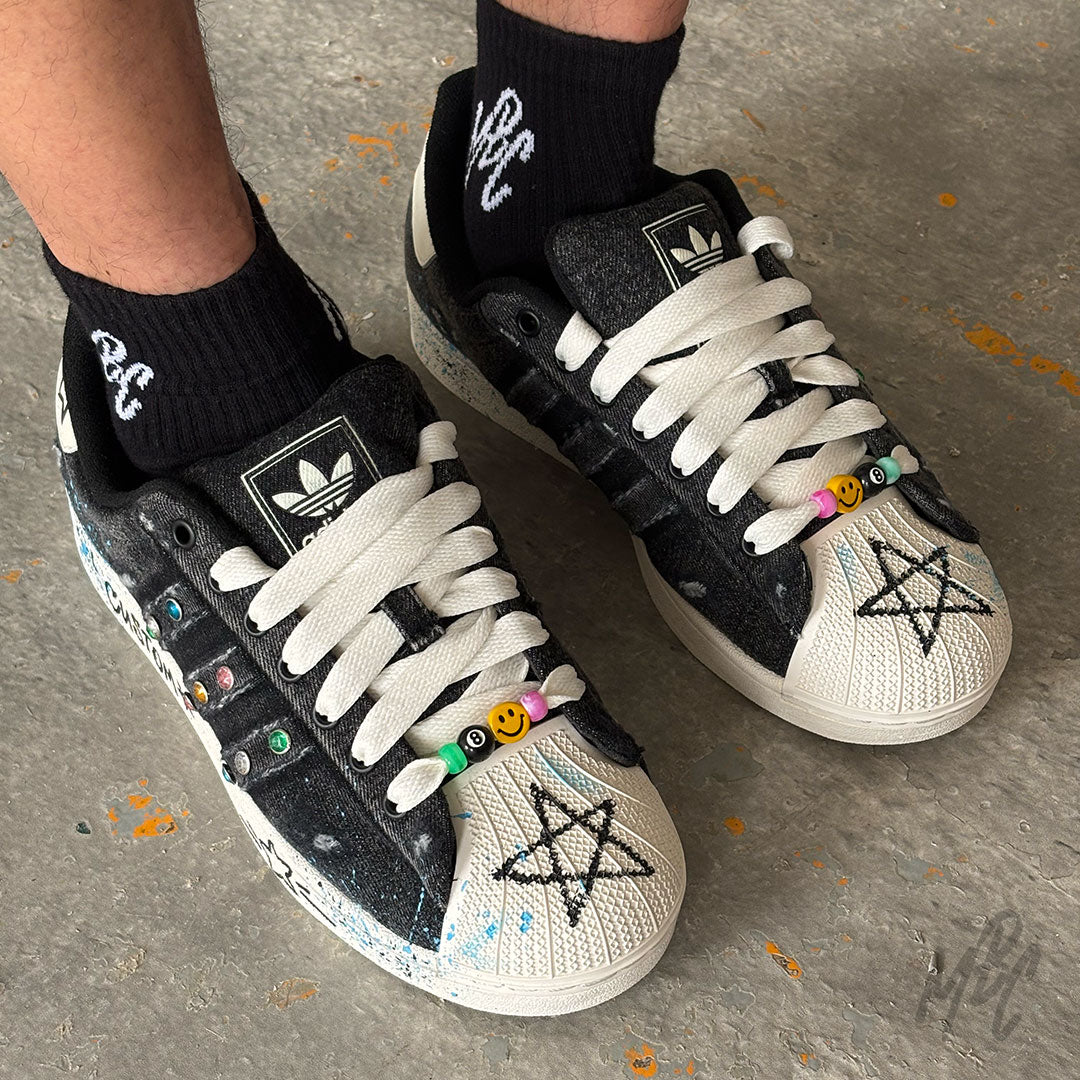 Top-down view of the Stardust Denim custom Adidas Superstar on foot, highlighting black denim uppers, colourful beaded lace accessory, crystal embellishments and hand-drawn star artwork.
