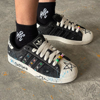 Side profile of the Stardust Denim custom Adidas Superstar on foot, showcasing black denim panels, colourful crystal detailing, white rope laces, blue paint splatters and hand-painted graphics.