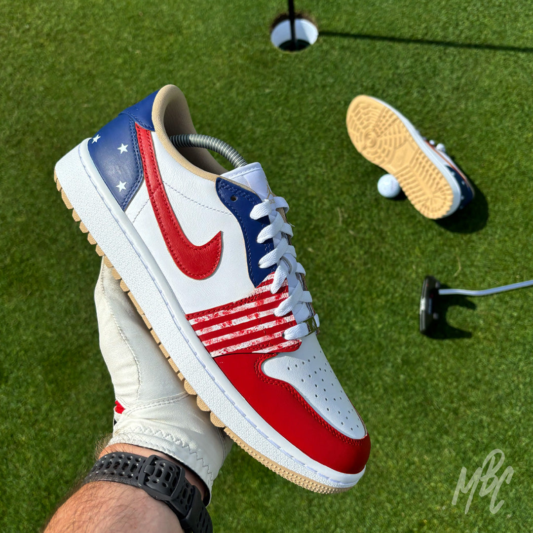 Stateside Drive custom Jordan 1 Low Golf shoes with hand-painted USA-inspired detailing, photographed on a golf course putting green beside the hole and flag.