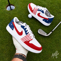 A pair of Nike Air Jordan 1 Low sneakers styled with an American flag theme, featuring a white leather base with red overlays on the toe, quarter, and Swoosh, and a blue star-patterned heel tab. The shoes are on a golf putting green. 