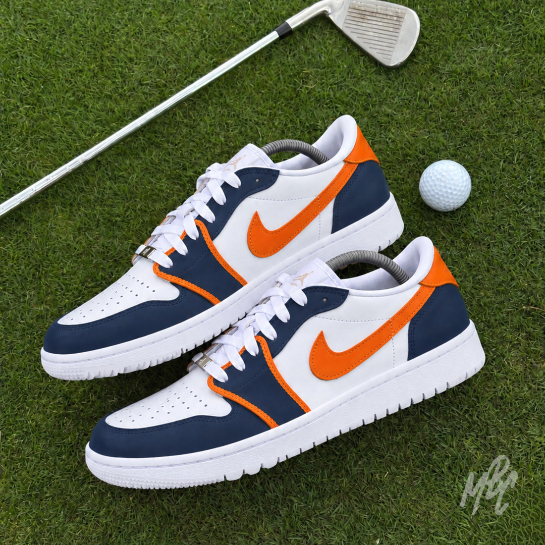 Two custom navy, white and orange golf shoes positioned on a putting green beside a golf ball and silver golf club.